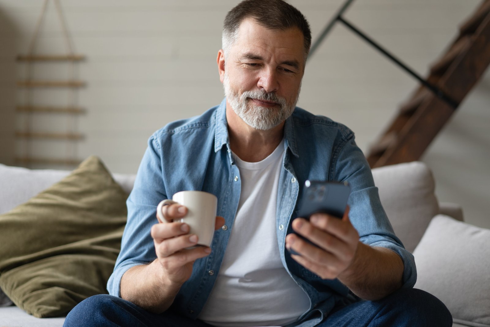 Happy smiling senior man using smartphone device while sitting on sofa at home. Mature man lying on couch reading messages on mobile phone, relaxing at home.