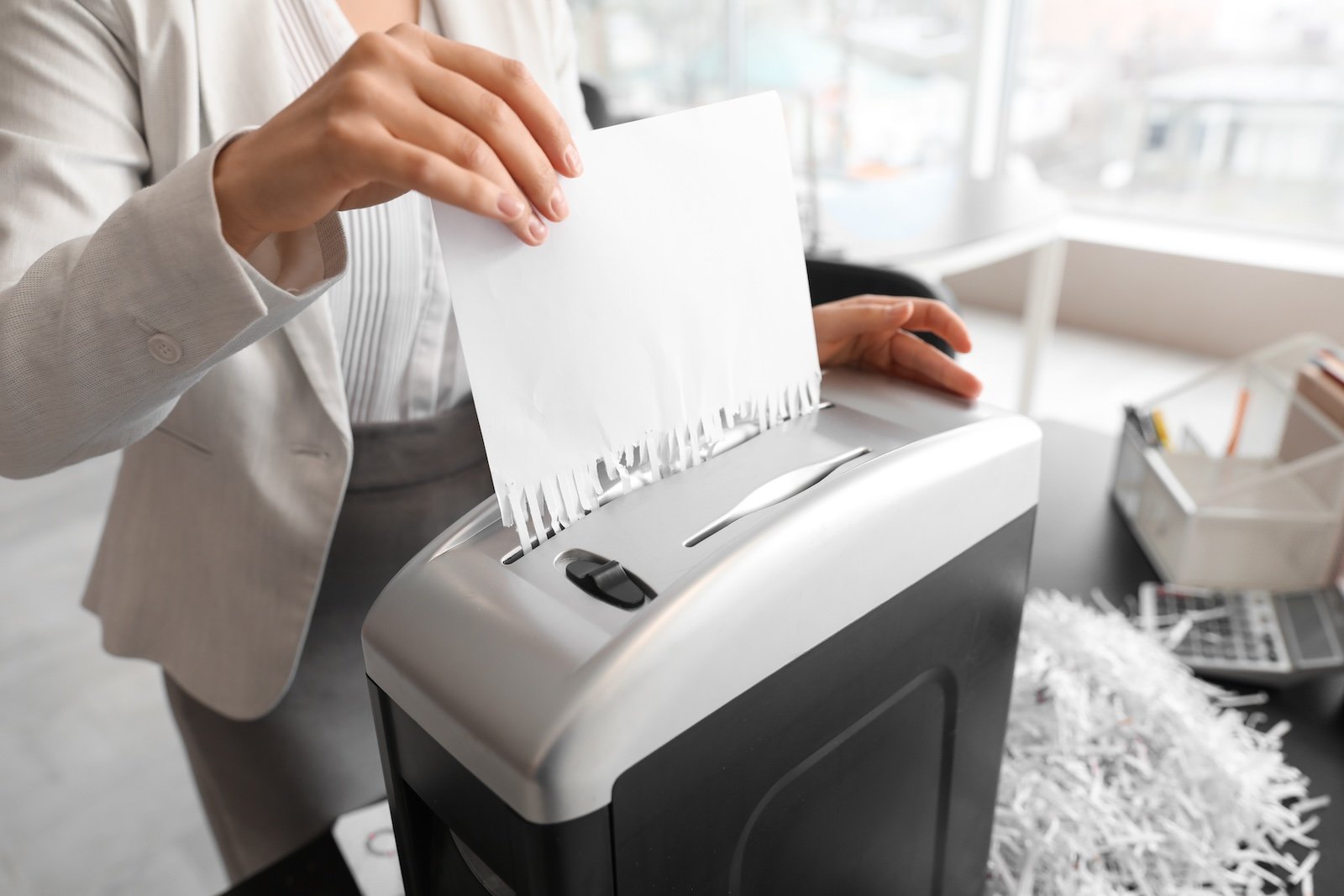 Young secretary destroying paper sheet using shredder in office, closeup